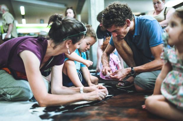Photographie d'une animation familiale où 2 parents et leur fils percent des pierres à l'aide d'outils en silex pour fabriquer des perles
