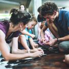 Photographie d'une animation familiale où 2 parents et leur fils percent des pierres à l'aide d'outils en silex pour fabriquer des perles