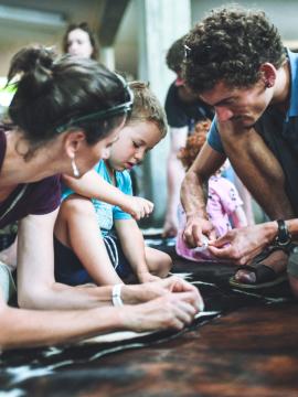 Photographie d'une animation familiale où 2 parents et leur fils percent des pierres à l'aide d'outils en silex pour fabriquer des perles