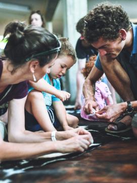 Photographie d'une animation familiale où 2 parents et leur fils percent des pierres à l'aide d'outils en silex pour fabriquer des perles