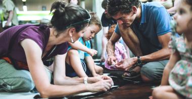Photographie d'une animation familiale où 2 parents et leur fils percent des pierres à l'aide d'outils en silex pour fabriquer des perles