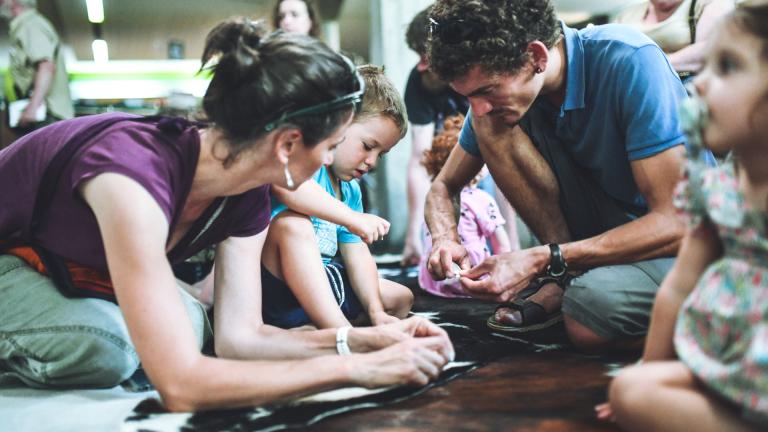 Photographie d'une animation familiale où 2 parents et leur fils percent des pierres à l'aide d'outils en silex pour fabriquer des perles
